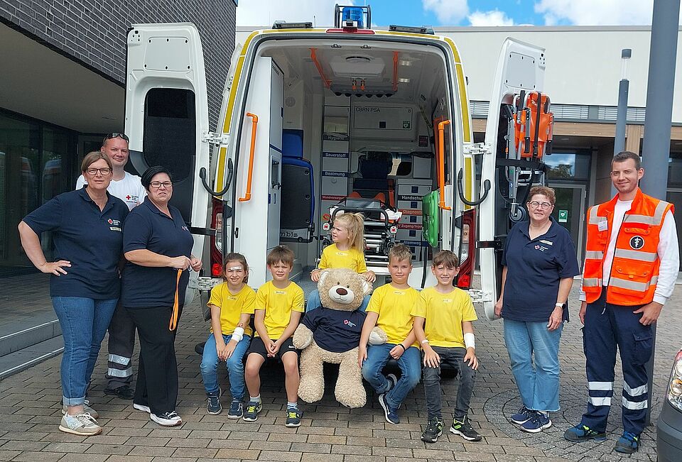 5 fröhliche Kinder sitzen vor dem Patientenraum des Krankenwagens. Rechts und links daneben freuen sich die Betreuende unseres Ortsvereines