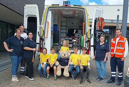 5 fröhliche Kinder sitzen vor dem Patientenraum des Krankenwagens. Rechts und links daneben freuen sich die Betreuende unseres Ortsvereines